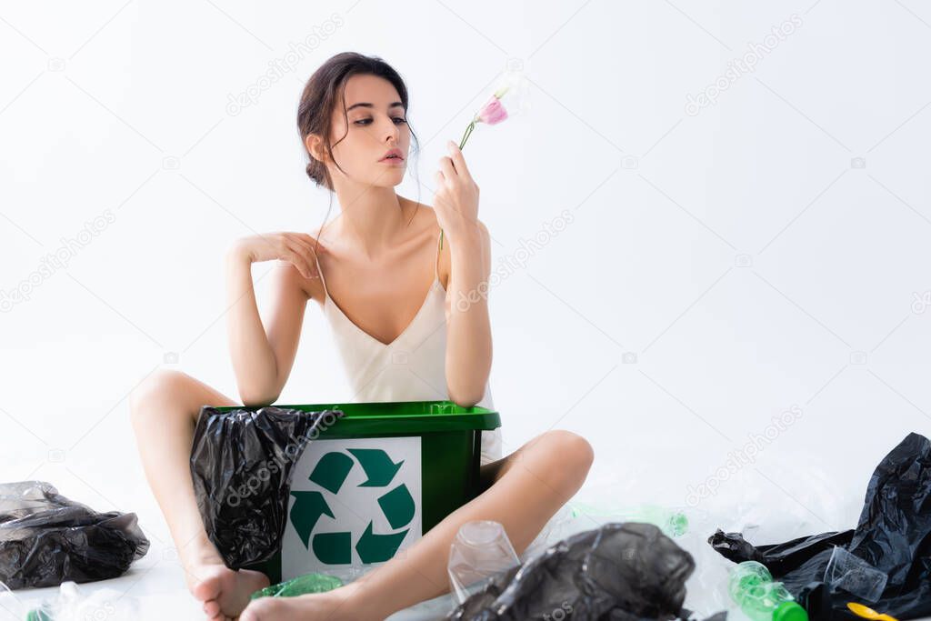 selective focus of young woman holding flower in wrap near plastic bags and trash can with recycle sign on white, ecology concept 