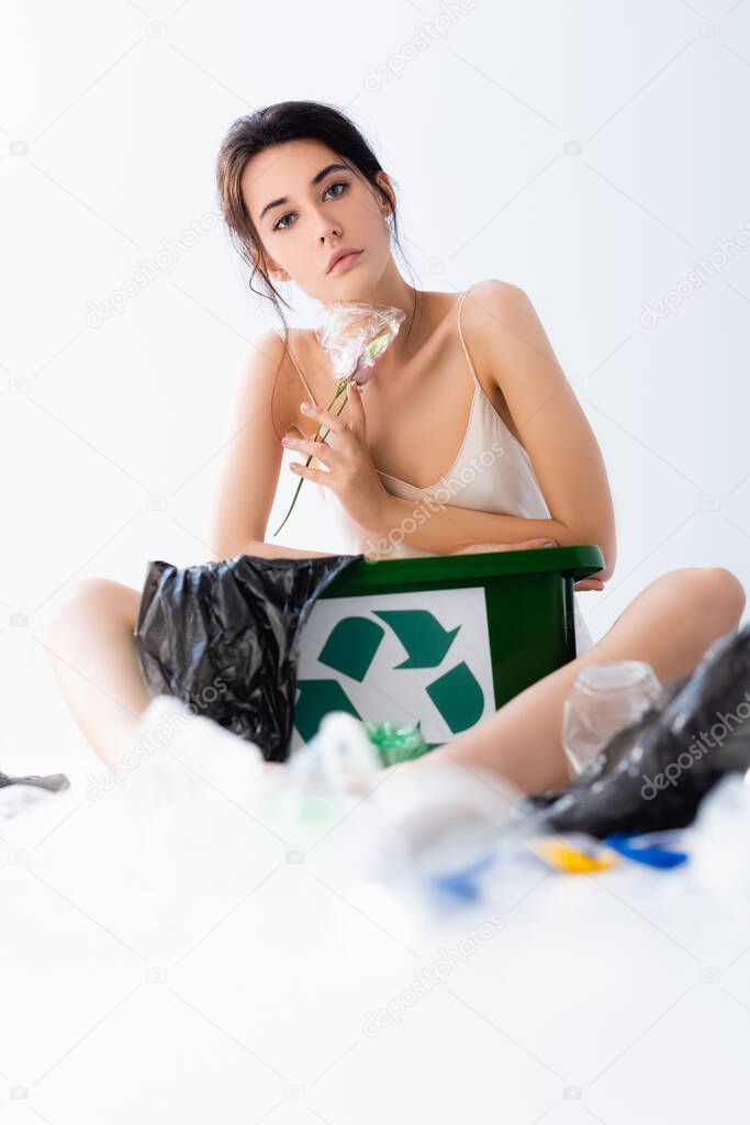 selective focus of young brunette woman holding flower in wrap near plastic bags and trash can with recycle sign on white, ecology concept 