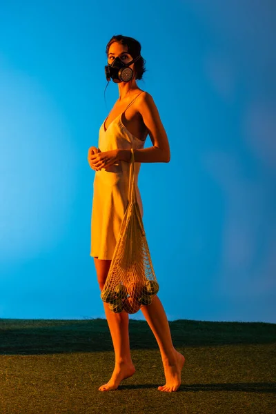 barefoot woman in gas mask and silk dress holding reusable string bag with fruits on blue