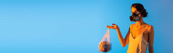horizontal image of woman in gas mask holding plastic bag with globe on blue