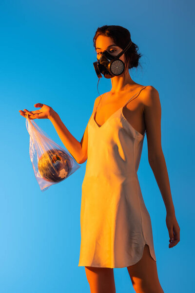 young woman in gas mask holding plastic bag with globe on blue, ecology concept 