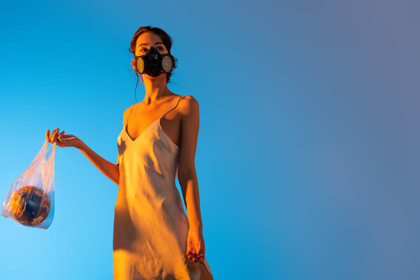 brunette woman in gas mask holding plastic bag with globe on blue, ecology concept 