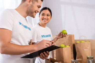 Seçici odak noktası Asyalı gönüllülerin yardım merkezinde panosu ve paketleri olan bir adamın yanında elma tutması. 