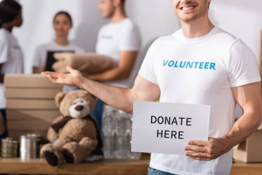 Selective focus of volunteer holding card with donate today lettering and pointing with hand in charity center  clipart