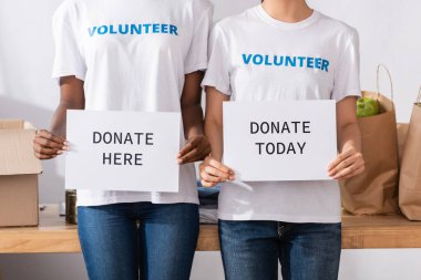 Cropped view of multiethnic volunteers holding card with donate here and today lettering in charity center  clipart