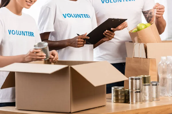 Vista Recortada Voluntarios Multiculturales Con Portapapeles Empacando Comida Mesa Centro — Foto de Stock