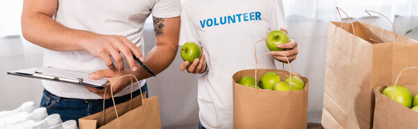 Panoramic crop of volunteer with clipboard pointing with hand near woman holding apples and packages in charity center 