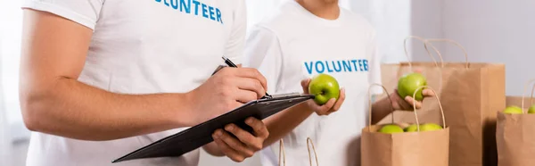 Foto Panorámica Voluntarios Escribiendo Portapapeles Sosteniendo Manzanas Cerca Paquetes Centro — Foto de Stock