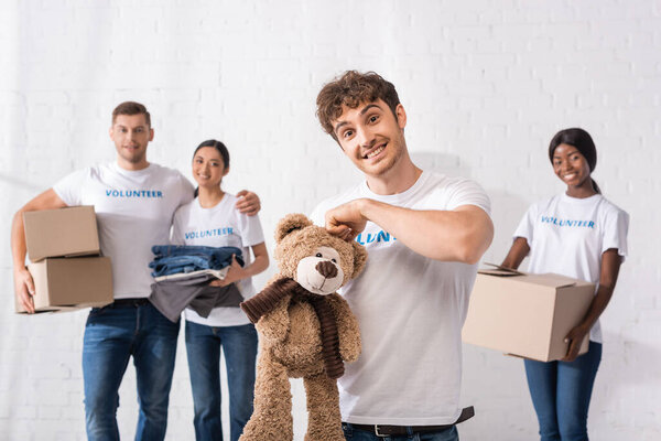 Селективный фокус человека с мягкой игрушкой, смотрящего в камеру рядом с многонациональными волонтерами с коробками на заднем плане 