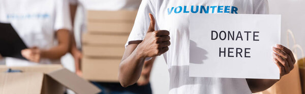 Панорамная ориентация африканского добровольца, показывающего большой палец и держащего карточку с надписью "Пожертвовать" 