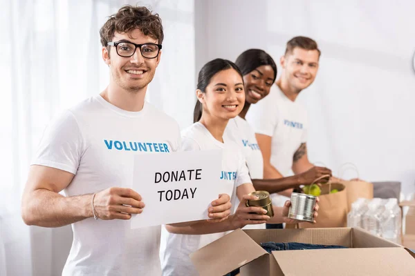 Selective focus of multicultural volunteers looking at camera while holding card with donate today lettering and donations in charity center