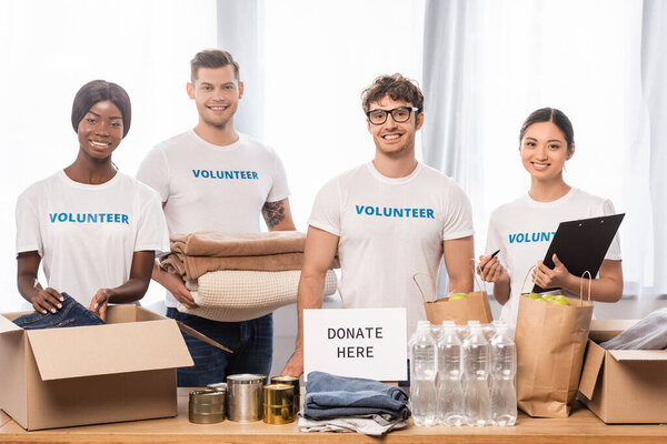 Multiethnic volunteers with clipboard and clothes standing near card with donate here lettering in charity center 
