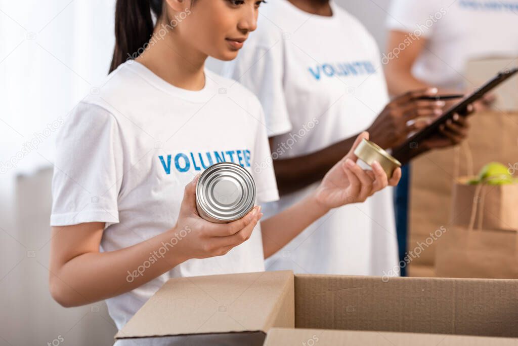 Selective focus of asian volunteer holding tin cans near cardboard box in charity center 
