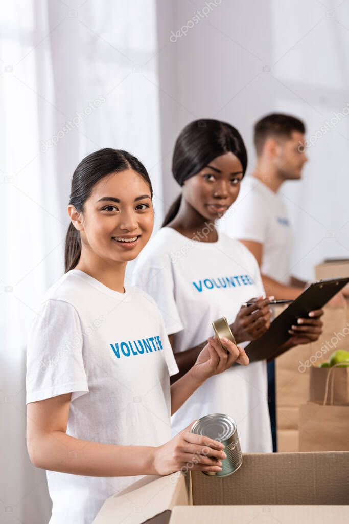Selective focus of asian volunteer with tin cans looking at camera near african american woman with clipboard in charity center 