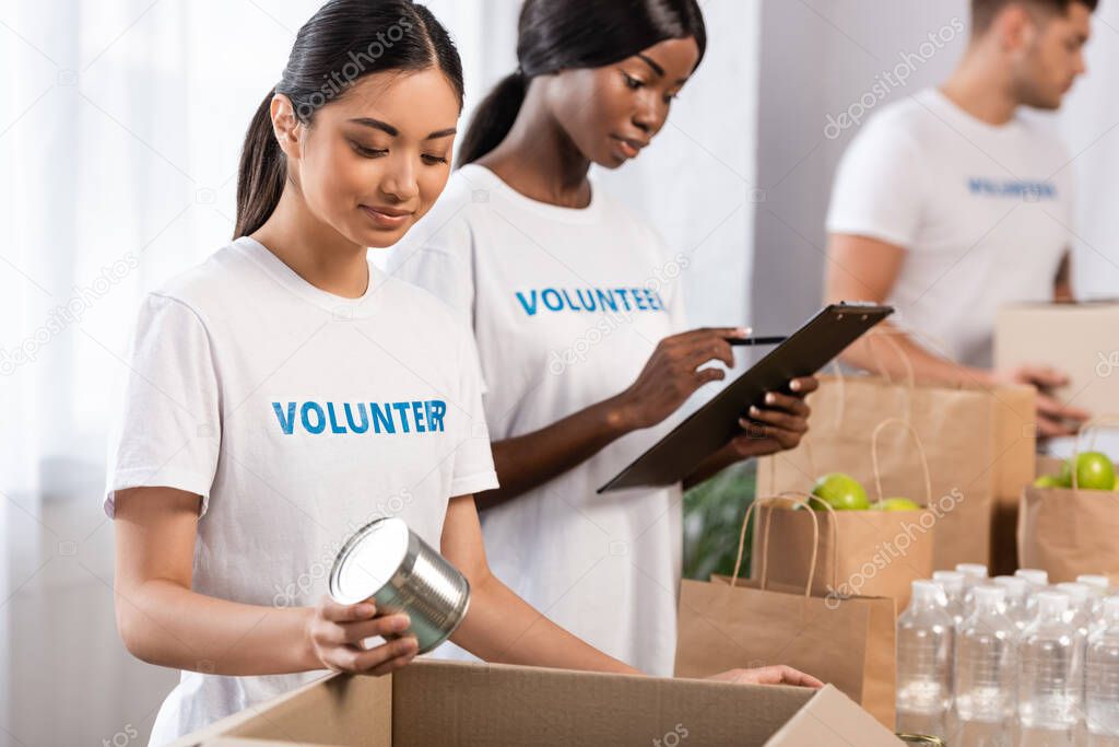 Selective focus of asian volunteer holding tin can near african american woman writing on clipboard in charity center 