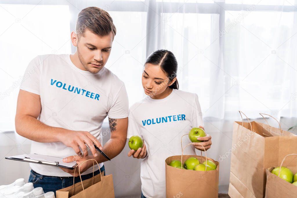 Selective focus of multicultural volunteers with clipboard and apples standing near packages in charity center 