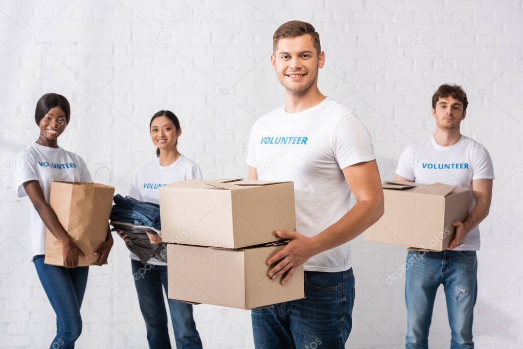 Selective focus of man holding carton boxes near volunteers with paper bag and clothes at background 