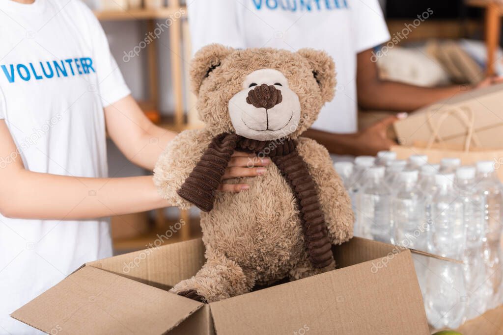 Cropped view of volunteer putting soft toy in carton box near packages with bottles of water in charity center 