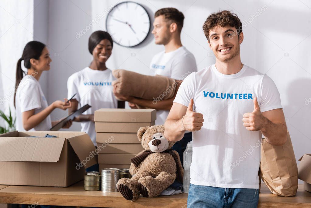 Selective focus of volunteer showing thumbs up near packages and multiethnic people in charity center