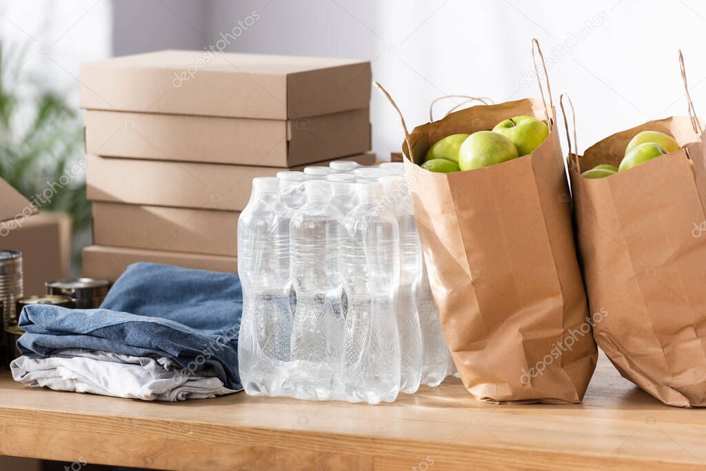 Paper bags with apples, bottles of water and clothes on table in charity center 