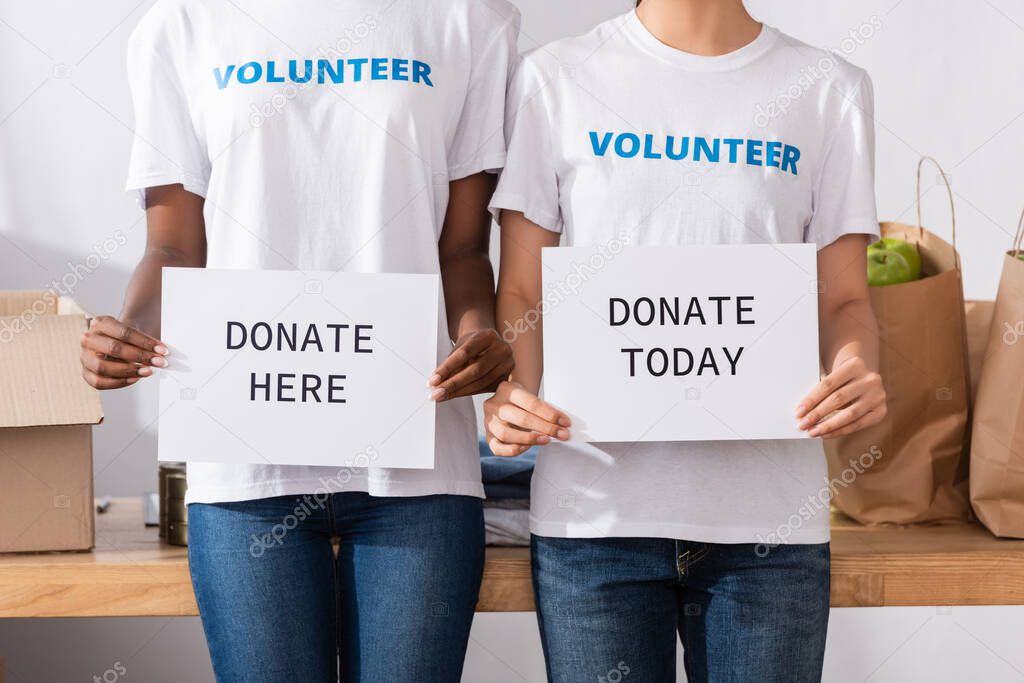 Cropped view of multiethnic volunteers holding card with donate here and today lettering in charity center 