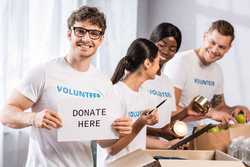 Selective focus of volunteer holding card with donate here lettering near multiethnic people in charity center 