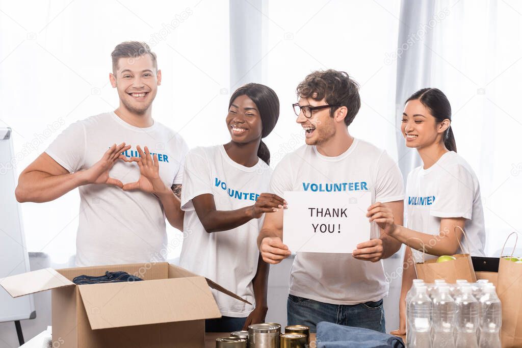 Selective focus of volunteer showing heart sign near laughing multiethnic people with lettering thank you on card and donations 