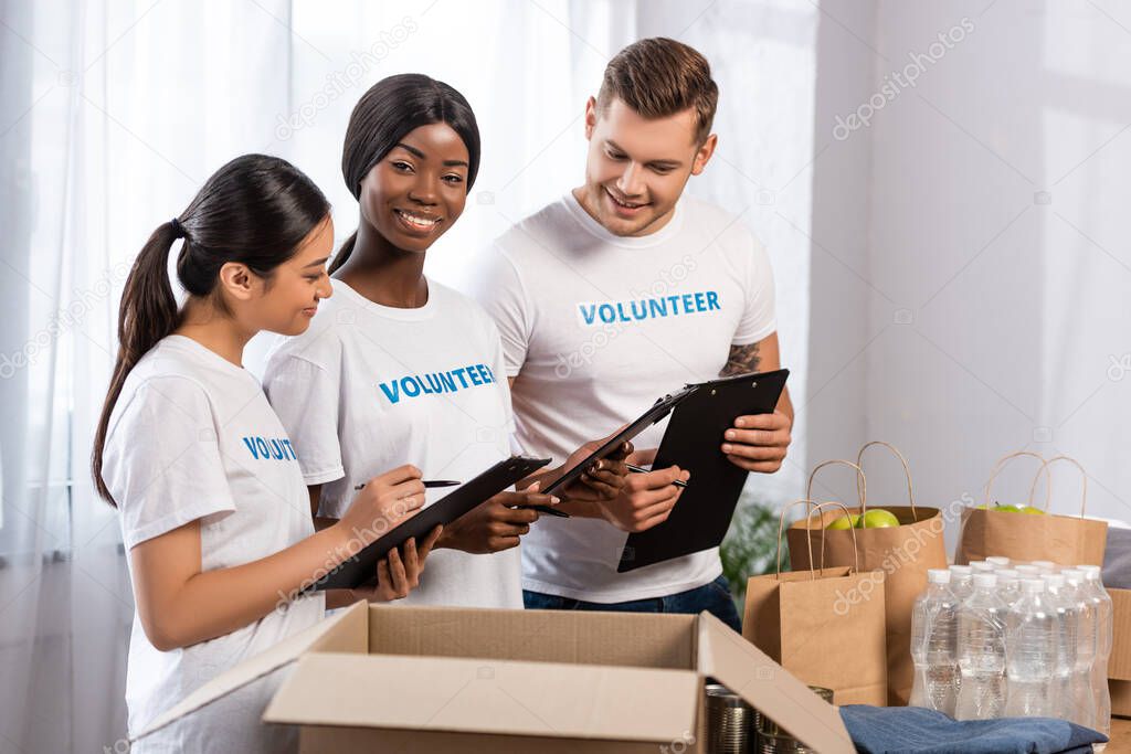 Selective focus of multiethnic volunteers with clipboards standing near donations in charity center 