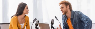young redhead broadcaster talking to brunette asian woman during interview, panoramic shot clipart