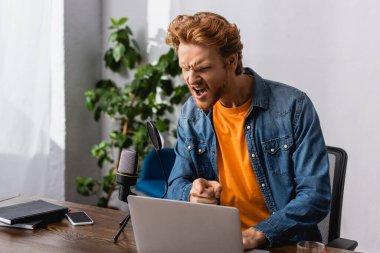 angry broadcaster in denim shirt pointing with finger while screaming in microphone near laptop clipart
