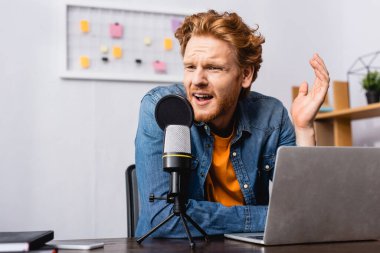 tense broadcaster in denim shirt gesturing while speaking in microphone near laptop clipart