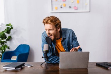 angry redhead announcer gesturing while screaming in microphone near laptop clipart
