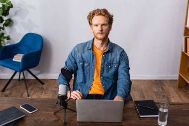 serious announcer in denim shirt sitting at workplace near microphone, gadgets and notebooks clipart