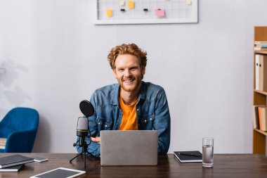 excited redhead announcer looking at camera near gadgets, microphone and notebooks clipart