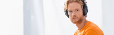 panoramic concept of serious redhead man in wireless headphones looking at camera clipart