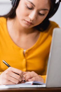 Laptop 'un yanında not defterine kablosuz kulaklık takan Asyalı genç tele-işçi seçici odağı