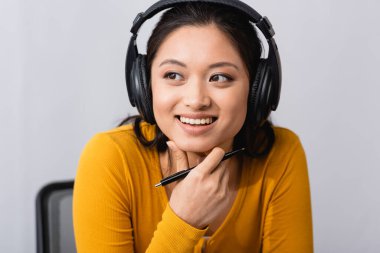 brunette asian woman in wireless headphones holding pen and touching chin while looking away clipart