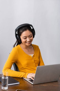 Esmer Asyalı, kablosuz kulaklıklı, evde dizüstü bilgisayarda yazan.