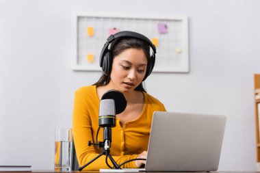 concentrated asian radio host in wireless headphones using laptop near microphone in studio clipart