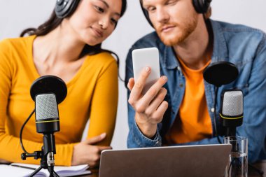Kablosuz kulaklıklarıyla Asyalı meslektaşının yanında akıllı telefon kullanan genç yayıncının seçici odak noktası