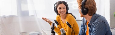 horizontal image of asian announcer in wireless headphones gesturing while talking to colleague in studio clipart