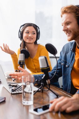 selective focus of asian announcer in wireless headphones gesturing near colleague in studio clipart
