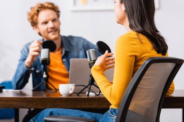 selective focus of redhead radio host near brunette woman talking and gesturing during interview clipart