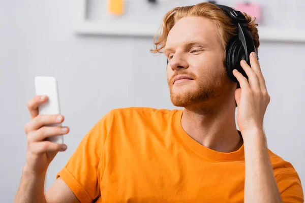 Selective Focus Dreamy Man Touching Wireless Headphones While Holding Smartphone — Stock Photo, Image