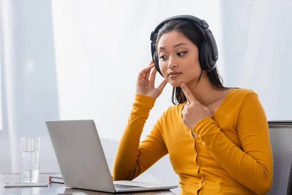 Curioso Freelancer Asiático Tocando Barbilla Auriculares Inalámbricos Mientras Mira Portátil — Foto de Stock