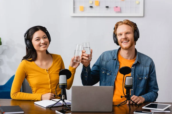 Interracial Couple Broadcasters Wireless Headphones Clinking Glasses Water While Looking — Stock Photo, Image