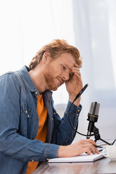 exhausted broadcaster touching head while sitting ner microphone, notebook and coffee cup