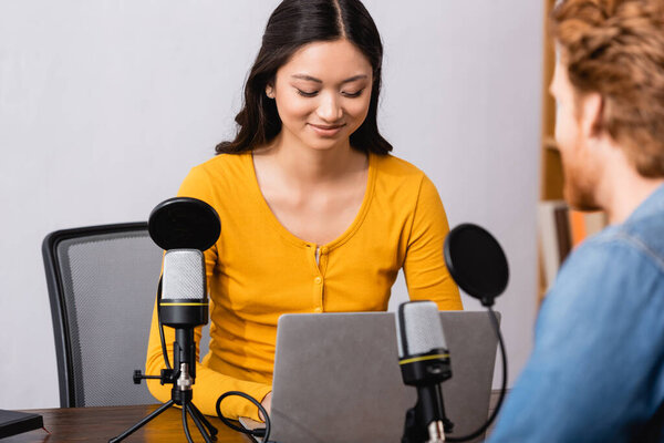 selective focus of brunette asian broadcaster using laptop during interview with man in radio studio