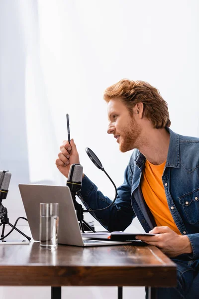 Jovem Emissora Ruiva Falando Perto Microfone Enquanto Segurando Caneta Notebook — Fotografia de Stock