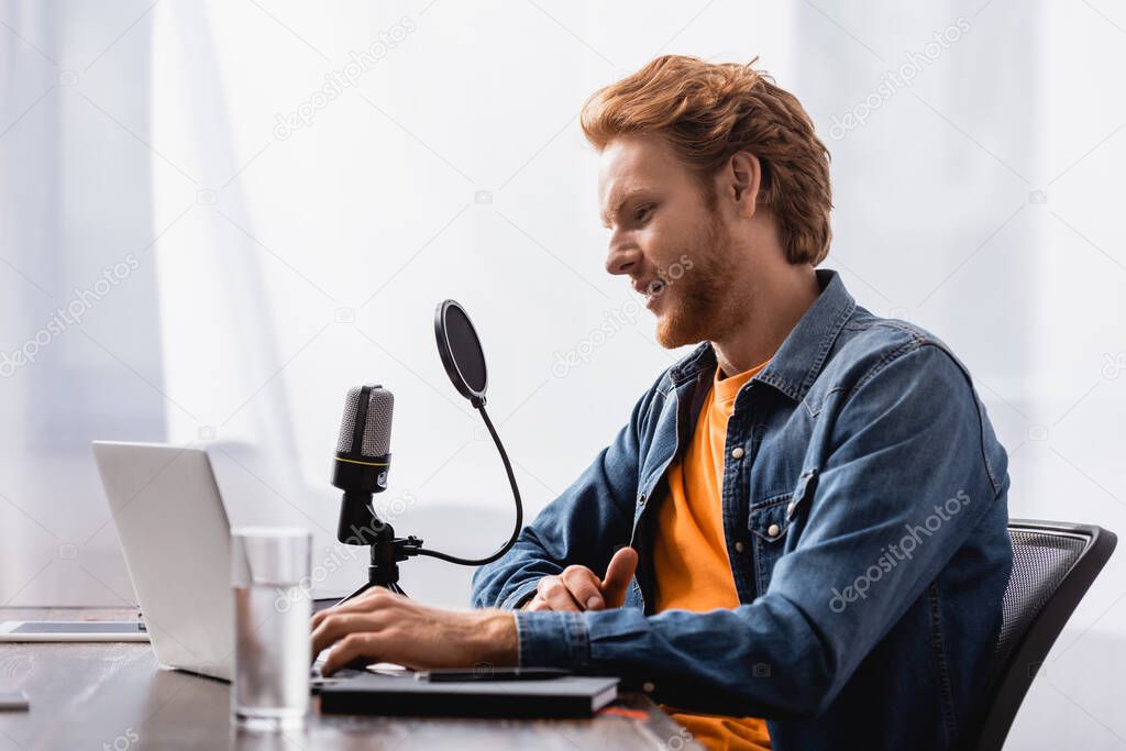 selective focus of redhead broadcaster talking in microphone while using laptop